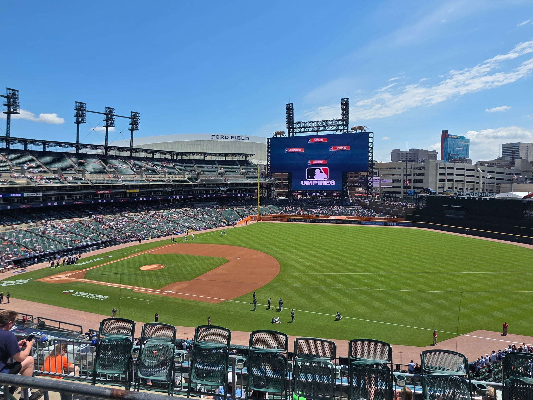 Wide view of Comerica Park in Detroit before a Seattle Mariners and Detroit Tigers game on a sunny afternoon, with the field, scoreboard and Ford Field visible in the background.
