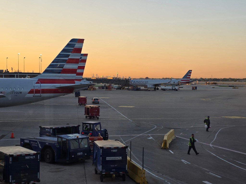 American Airlines planes sit at airport gates as the sun sets behind the terminal.