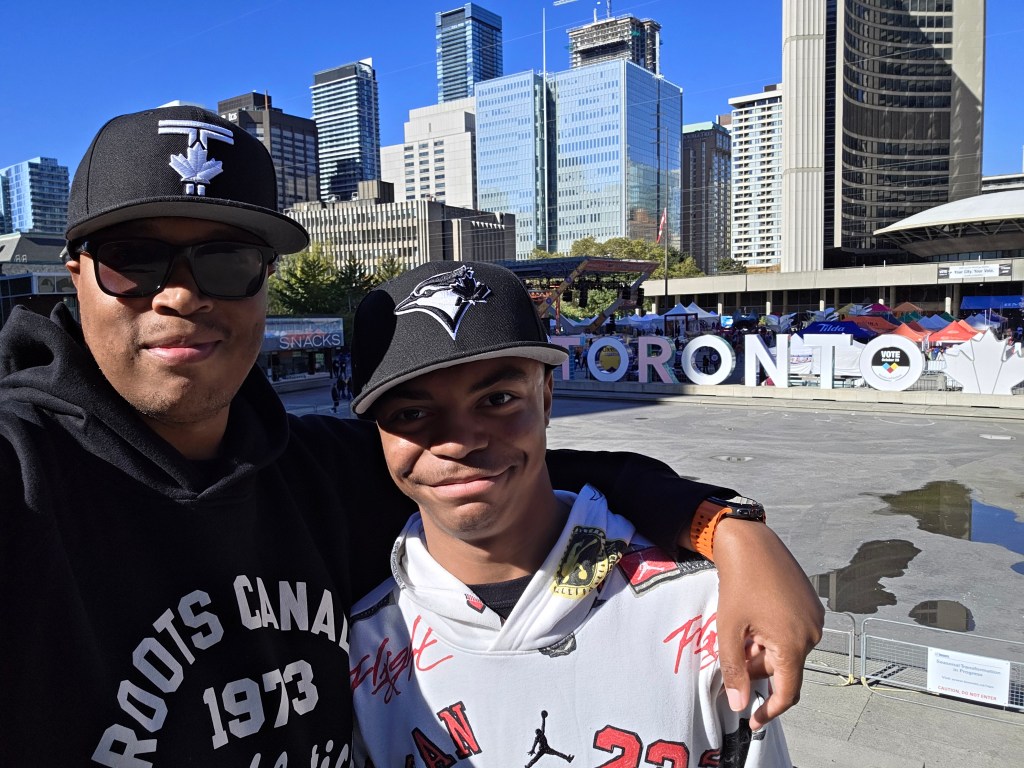 Father and teenage son smiling for a selfie in front of the Toronto sign at Nathan Phillips Square in downtown Toronto, Canada.
